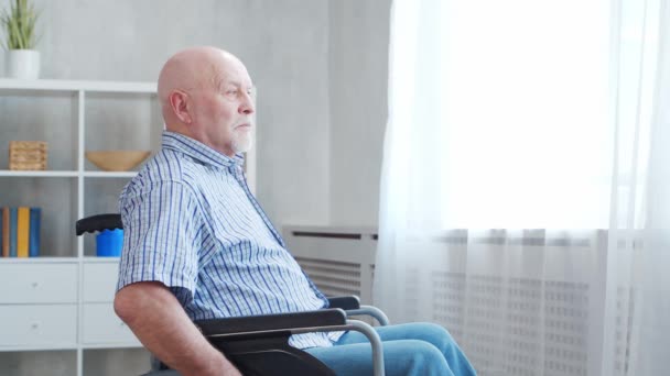 Le vieil homme handicapé est assis seul dans un fauteuil roulant à la maison. Une personne handicapée en fauteuil roulant regarde par la fenêtre. Paralysie et invalidité. — Video