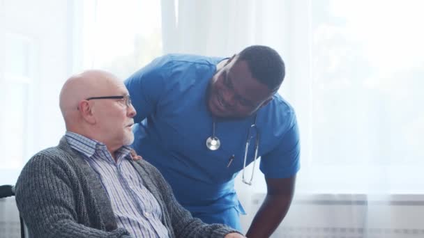 African-American caregiver and old disabled man in a wheelchair. Professional nurse and handicapped patient in a nursing home. Assistance, rehabilitation and health care. — Stock Video