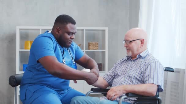 Afro-Amerikaanse verzorger meet de gehandicapte oude man bloeddruk. Professionele verpleegkundige en gehandicapte patiënt in een verpleeghuis. Bijstand, rehabilitatie en gezondheidszorg. — Stockvideo