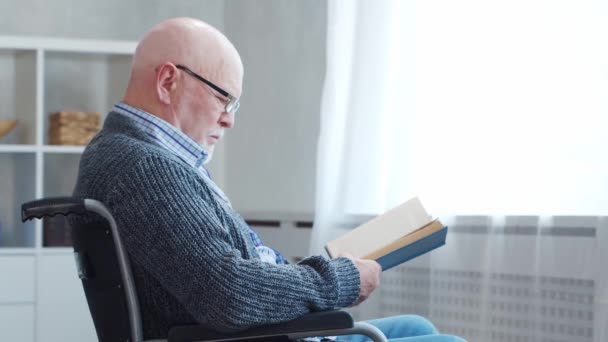 A rokkant öregember egyedül ül otthon egy tolószékben. Egy kerekesszékes mozgássérült olvassa a könyvet. Bénulás és fogyatékosság. — Stock videók