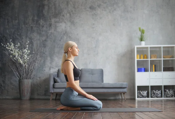 Chica joven y deportiva en ropa deportiva está haciendo ejercicios de yoga en el interior del hogar. Ajuste y delgada mujer rubia entra en los deportes y la aptitud. Salud, quemaduras de grasa y bienestar. — Foto de Stock