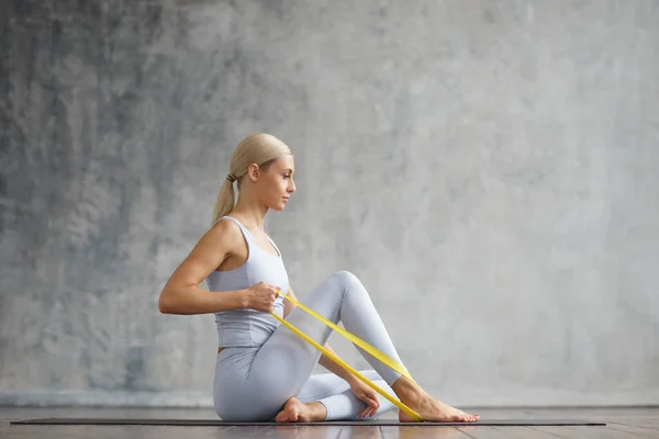 Young and sporty girl in sportswear is doing exercises in home interior using resistance band. Fit and slender blond woman goes in for sports and fitness. Healthcare, fat burn and wellness. — Stock Photo, Image