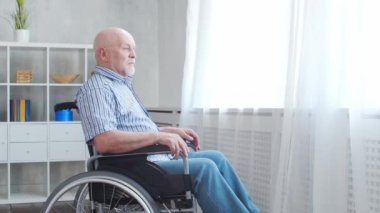 Engelli yaşlı adam evde tek başına tekerlekli sandalyede oturuyor. Tekerlekli sandalyedeki engelli pencereden dışarı bakıyor. Felç ve sakatlık.