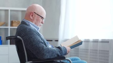 Engelli yaşlı adam evde tek başına tekerlekli sandalyede oturuyor. Tekerlekli sandalyedeki bir özürlü kitabı okuyor. Felç ve sakatlık.