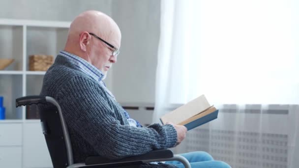 Disabled old man is sitting in a wheelchair at home alone. A handicapped person in a wheelchair is reading the book. Paralysis and disability. — Stock Video