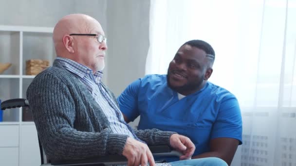 Afro-Amerikaanse verzorger en oude gehandicapte man in een rolstoel. Professionele verpleegkundige en gehandicapte patiënt in een verpleeghuis. Bijstand, rehabilitatie en gezondheidszorg. — Stockvideo