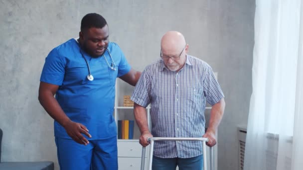 Afro-Amerikaanse verzorger leert gehandicapte oude man lopen met Walker. Professionele verpleegkundige en gehandicapte patiënt in een verpleeghuis. Bijstand, rehabilitatie en gezondheidszorg. — Stockvideo