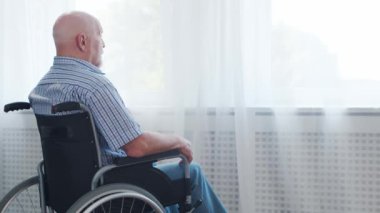 Engelli yaşlı adam evde tek başına tekerlekli sandalyede oturuyor. Tekerlekli sandalyedeki engelli pencereden dışarı bakıyor. Felç ve sakatlık.