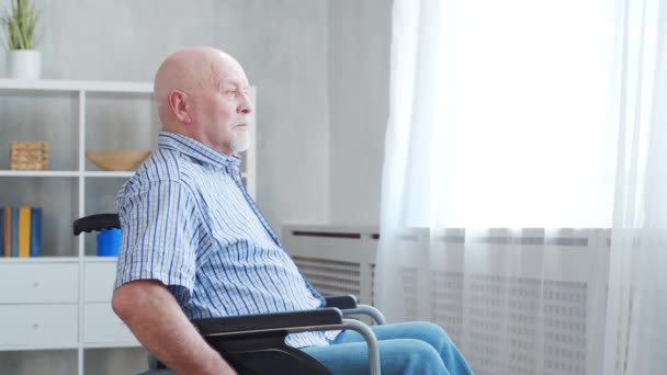 Le vieil homme handicapé est assis seul dans un fauteuil roulant à la maison. Une personne handicapée en fauteuil roulant regarde par la fenêtre. Paralysie et invalidité. — Video