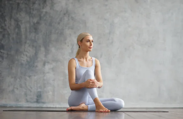 Jong en sportief meisje in sportkleding doet yoga oefeningen in het interieur. Fit en slanke blonde vrouw gaat in voor sport en fitness. Gezondheidszorg, vetverbranding en welzijn. — Stockfoto