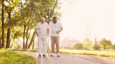 Afro-Amerikalı bakıcı engelli ihtiyara Walker 'la yürümeyi öğretiyor. Parktaki profesyonel hemşire ve özürlü hasta. Yardım, rehabilitasyon ve sağlık hizmetleri. Altın saat gün batımı.