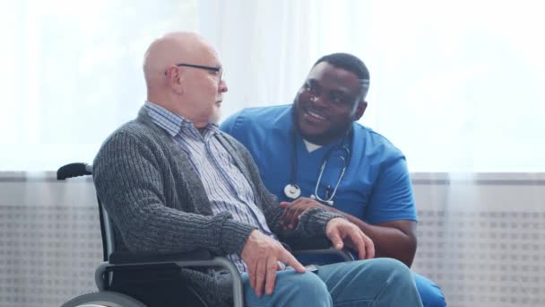 Afro-Amerikaanse verzorger en oude gehandicapte man in een rolstoel. Professionele verpleegkundige en gehandicapte patiënt in een verpleeghuis. Bijstand, rehabilitatie en gezondheidszorg. — Stockvideo