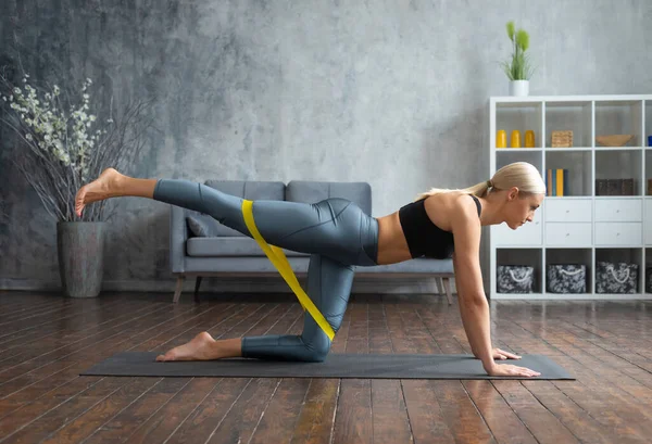 Menina jovem e desportiva em sportswear está fazendo exercícios em casa interior usando banda de resistência. Fit e mulher loira esbelta entra para esportes e fitness. Saúde, queima de gordura e bem-estar. — Fotografia de Stock