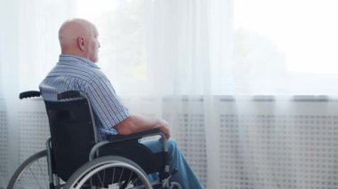 Engelli yaşlı adam evde tek başına tekerlekli sandalyede oturuyor. Tekerlekli sandalyedeki engelli pencereden dışarı bakıyor. Felç ve sakatlık.