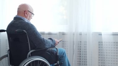 Engelli yaşlı adam evde tek başına tekerlekli sandalyede oturuyor. Tekerlekli sandalyedeki bir özürlü kitabı okuyor. Felç ve sakatlık.