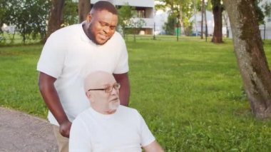 Afro-Amerikalı bakıcı ve tekerlekli sandalyedeki yaşlı engelli adam. Parktaki profesyonel hemşire ve özürlü hasta. Yardım, rehabilitasyon ve sağlık hizmetleri.