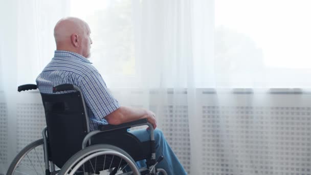 El viejo discapacitado está sentado en una silla de ruedas en casa solo. Una persona discapacitada en silla de ruedas está mirando por la ventana. Parálisis y discapacidad. — Vídeos de Stock