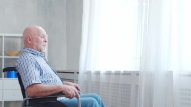 Engelli yaşlı adam evde tek başına tekerlekli sandalyede oturuyor. Tekerlekli sandalyedeki engelli pencereden dışarı bakıyor. Felç ve sakatlık.