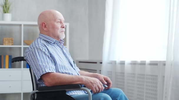 Ein behinderter alter Mann sitzt allein zu Hause im Rollstuhl. Ein Behinderter im Rollstuhl schaut aus dem Fenster. Lähmung und Behinderung. — Stockvideo