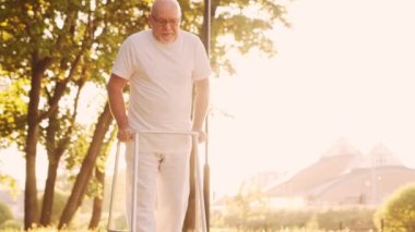 Engelli yaşlı adam bir yürüteçle yürüyor. Parktaki özürlü hasta. Yardım, rehabilitasyon ve sağlık hizmetleri. Altın saat gün batımı.