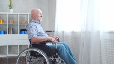 Engelli yaşlı adam evde tek başına tekerlekli sandalyede oturuyor. Tekerlekli sandalyedeki engelli pencereden dışarı bakıyor. Felç ve sakatlık.