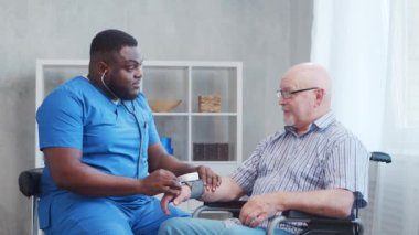 Afro-Amerikalı bakıcı engelli yaşlı adamın kan basıncını ölçüyor. Bakımevindeki profesyonel hemşire ve özürlü hasta. Yardım, rehabilitasyon ve sağlık hizmetleri.