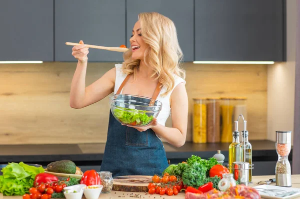 Jonge vrouw bereidt eten in de moderne keuken. Mooi blond meisje kookt thuis salade. Vegetarische gezonde voedselbereiding. — Stockfoto