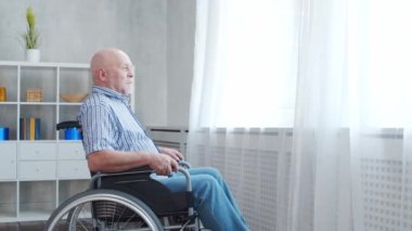 Engelli yaşlı adam evde tek başına tekerlekli sandalyede oturuyor. Tekerlekli sandalyedeki engelli pencereden dışarı bakıyor. Felç ve sakatlık.