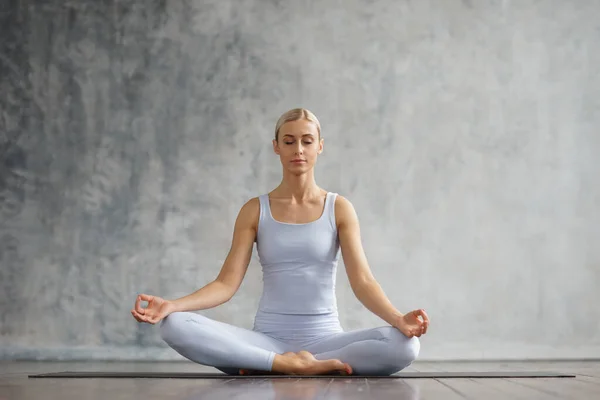 Jong en sportief meisje in sportkleding doet yoga oefeningen in het interieur. Fit en slanke blonde vrouw gaat in voor sport en fitness. Gezondheidszorg, vetverbranding en welzijn. — Stockfoto