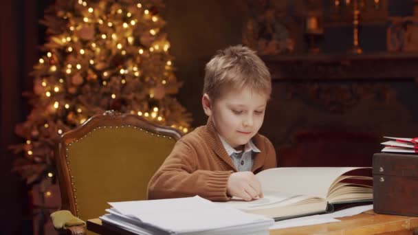 O rapazinho lê um livro de magia enquanto está sentado à mesa. Interior da casa com árvore de Natal e lareira. Conceito de Natal. — Vídeo de Stock