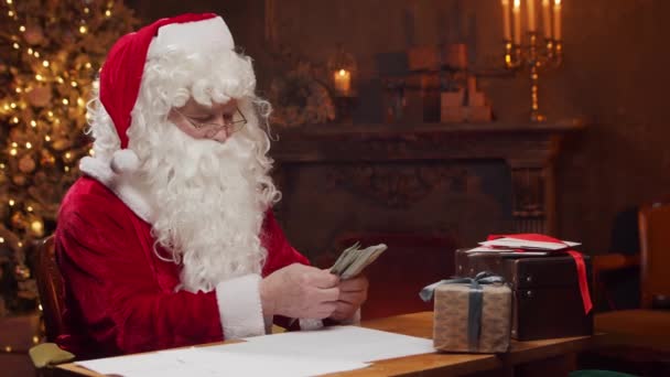 Lugar de trabajo de Santa Claus. Alegre Santa está contando dinero mientras está sentado en la mesa. Chimenea y árbol de Navidad en el fondo. Concepto de Navidad. — Vídeos de Stock