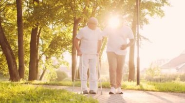 Afro-Amerikalı bakıcı engelli ihtiyara Walker 'la yürümeyi öğretiyor. Parktaki profesyonel hemşire ve özürlü hasta. Yardım, rehabilitasyon ve sağlık hizmetleri. Altın saat gün batımı.