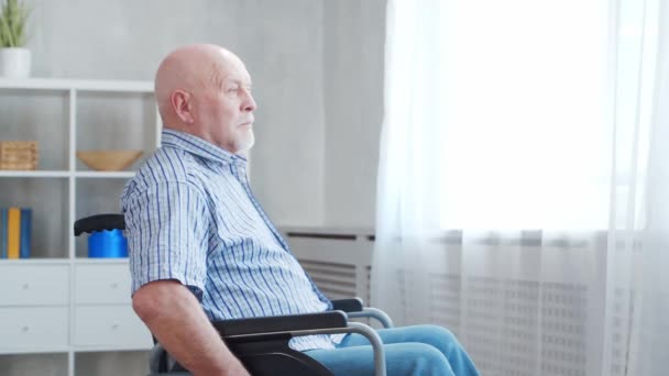 Il vecchio disabile è seduto su una sedia a rotelle a casa da solo. Una persona handicappata su una sedia a rotelle guarda fuori dalla finestra. Paralisi e disabilità. — Video Stock