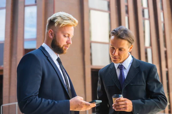 Un homme d'affaires confiant et son collègue utilisent un smartphone devant un immeuble de bureaux moderne. Les investisseurs financiers parlent en plein air. Banques et entreprises. — Photo