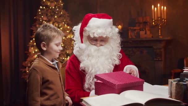 Babbo Natale e il bambino. Allegro Babbo Natale sta lavorando mentre è seduto a tavola. Camino e Albero di Natale sullo sfondo. Concetto Natale. — Video Stock