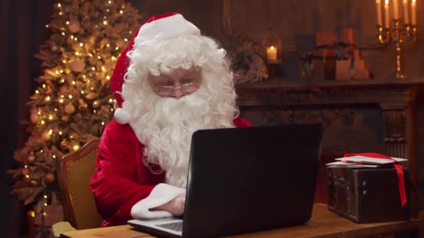 Arbeitsplatz des Weihnachtsmannes. Der fröhliche Weihnachtsmann arbeitet am Laptop, während er am Tisch sitzt. Kamin und Weihnachtsbaum im Hintergrund. Weihnachtskonzept. — Stockvideo
