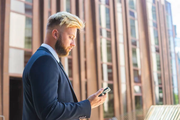 Homme d'affaires confiant devant un immeuble de bureaux moderne. Investisseur financier utilise smartphone en plein air. Banques et entreprises. — Photo