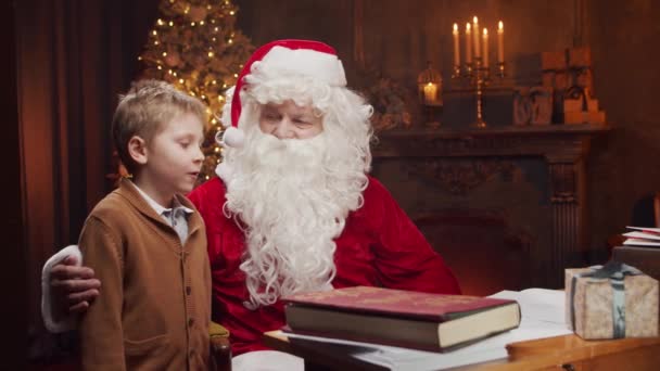 Santa Claus y el niño pequeño. Alegre Santa está trabajando mientras está sentado a la mesa. Chimenea y árbol de Navidad en el fondo. Concepto de Navidad. — Vídeo de stock