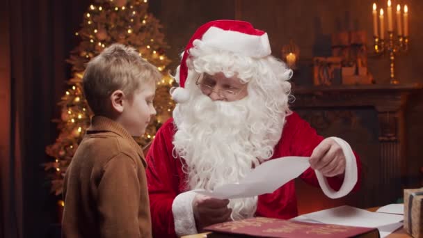 Santa Claus y el niño pequeño. Alegre Santa está trabajando mientras está sentado a la mesa. Chimenea y árbol de Navidad en el fondo. Concepto de Navidad. — Vídeos de Stock