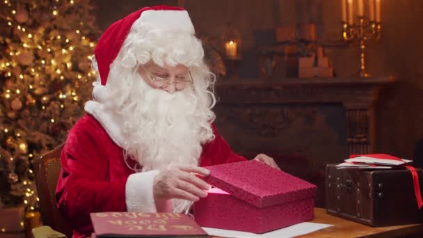 圣诞老人的工作场所。愉快的圣诞老人坐在餐桌前，一边在礼品盒上做鬼脸。背景是萤火虫和圣诞树。圣诞节的概念. — 图库视频影像