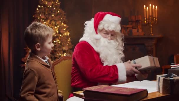 Santa Claus y el niño pequeño. Alegre Santa está trabajando mientras está sentado a la mesa. Chimenea y árbol de Navidad en el fondo. Concepto de Navidad. — Vídeos de Stock