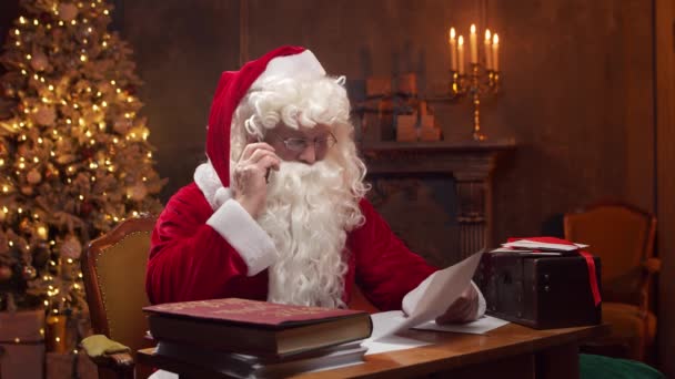 Lugar de trabajo de Santa Claus. Alegre Santa está trabajando con el teléfono inteligente mientras está sentado en la mesa. Chimenea y árbol de Navidad en el fondo. Concepto de Navidad. — Vídeos de Stock