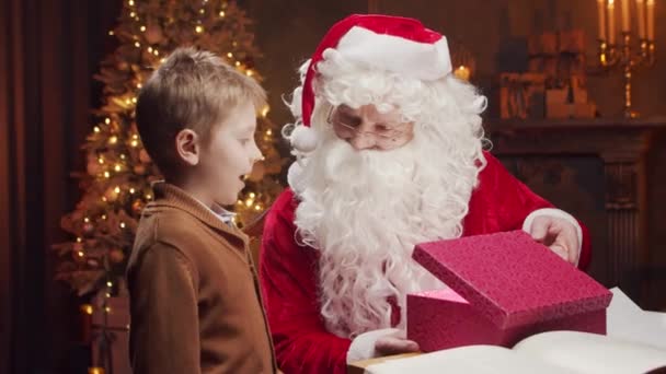 Weihnachtsmann und kleiner Junge. Der fröhliche Weihnachtsmann sitzt am Tisch und arbeitet. Kamin und Weihnachtsbaum im Hintergrund. Weihnachtskonzept. — Stockvideo