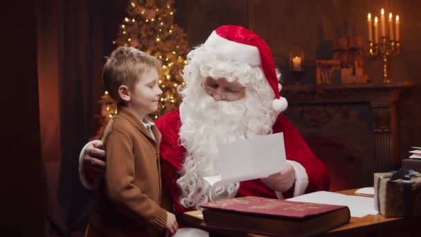 サンタクロースと男の子。陽気サンタはテーブルに座っている間に働いています。背景にある暖炉とクリスマスツリー。クリスマスのコンセプト. — ストック動画