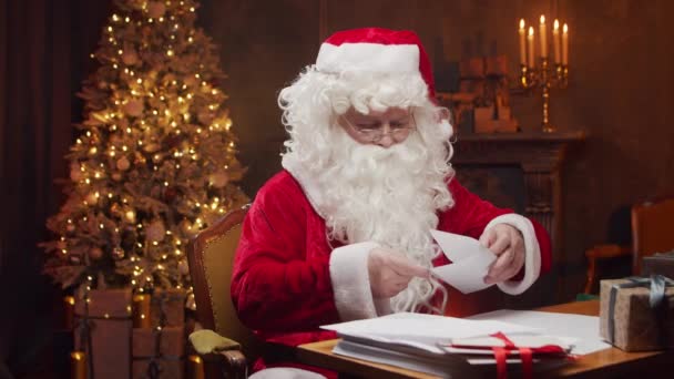 サンタクロースの職場。陽気サンタはテーブルに座っている間に子供たちからの手紙を読んでいます。背景にある暖炉とクリスマスツリー。クリスマスのコンセプト. — ストック動画