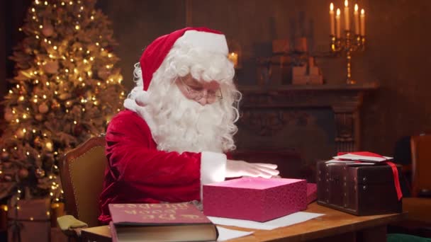 Lugar de trabajo de Santa Claus. Alegre Santa está conjurando sobre la caja de regalo mientras está sentado a la mesa. Chimenea y árbol de Navidad en el fondo. Concepto de Navidad. — Vídeos de Stock