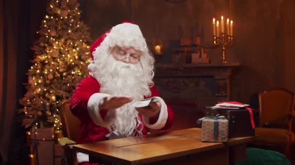 Lugar de trabajo de Santa Claus. Alegre Santa está contando dinero mientras está sentado en la mesa. Chimenea y árbol de Navidad en el fondo. Concepto de Navidad. — Vídeos de Stock