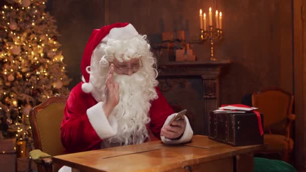 Lugar de trabajo de Santa Claus. Alegre Santa está trabajando con el teléfono inteligente mientras está sentado en la mesa. Chimenea y árbol de Navidad en el fondo. Concepto de Navidad. — Vídeos de Stock