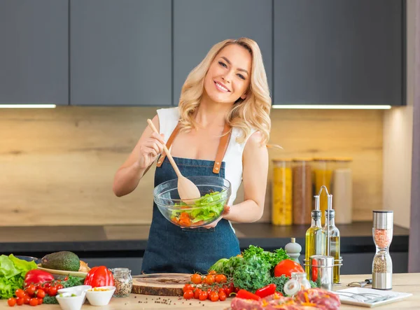 Young woman prepares food on modern kitchen. Beautiful blond girl is cooking salad at home. Vegetarian healthy food preparation.