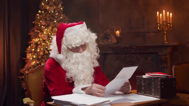 Luogo di lavoro di Babbo Natale. Babbo Natale allegro sta leggendo le lettere dei bambini mentre siede al tavolo. Camino e Albero di Natale sullo sfondo. Concetto Natale. — Video Stock